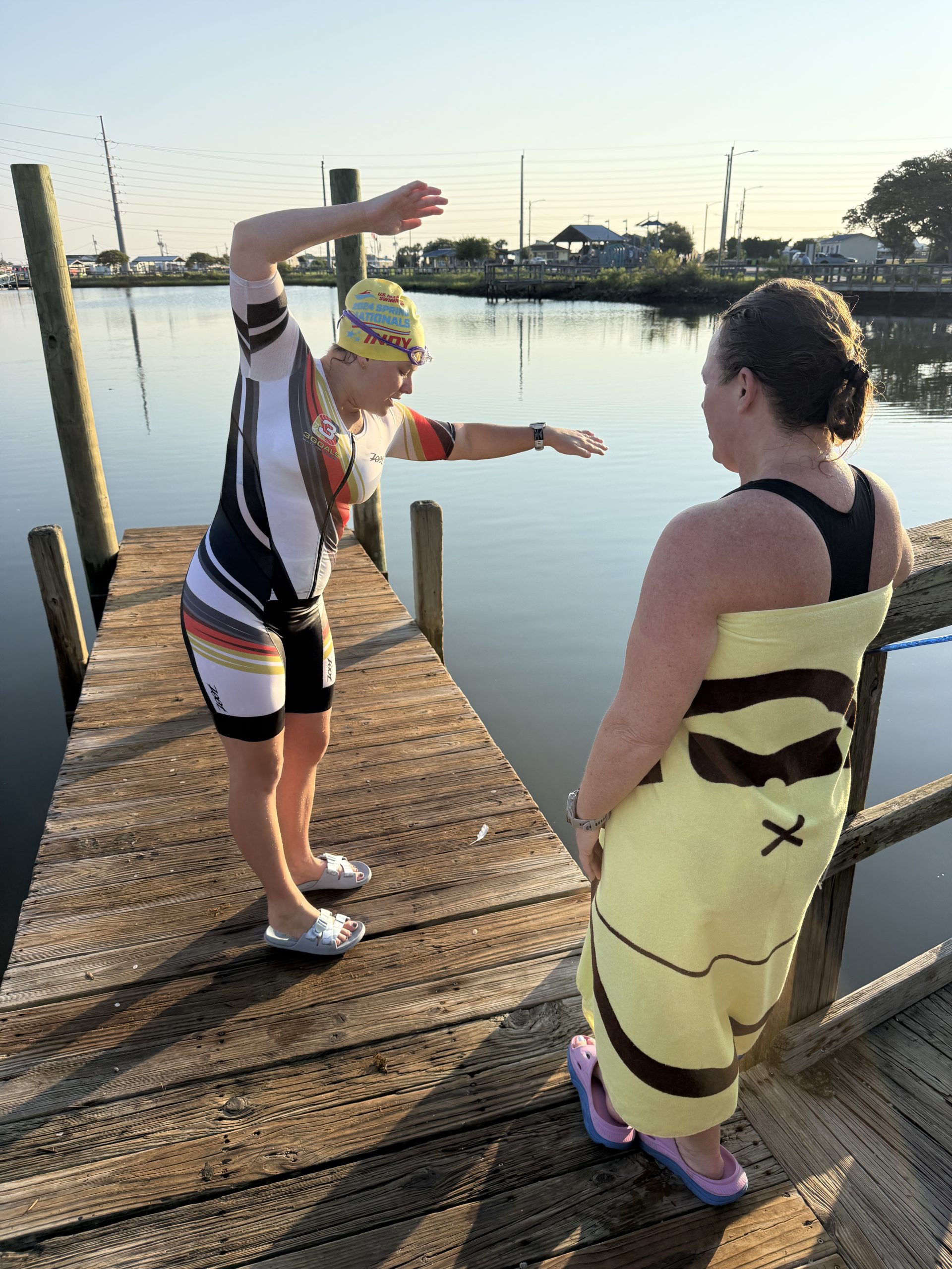 A Team 3G athlete wearing a swim cap demonstrates an arm movement beside a teammate on a dock — join Team 3G.