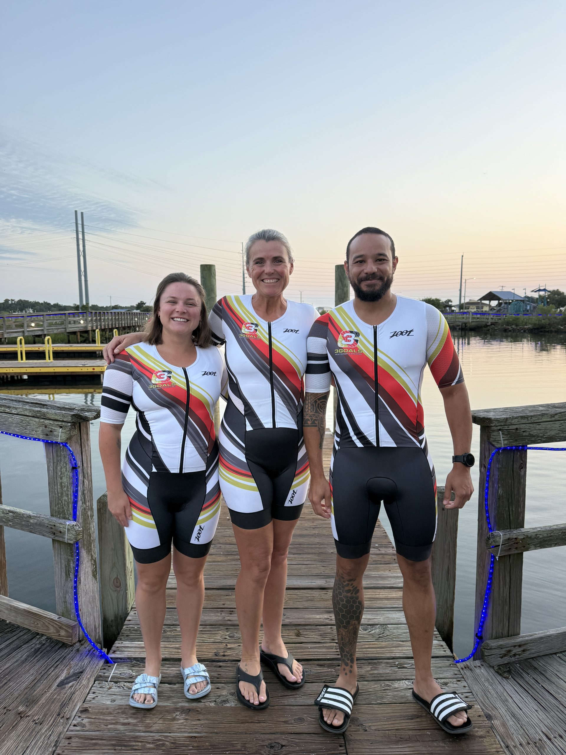 Three Team 3G teammates in matching triathlon kits on a waterfront dock — join Team 3G.